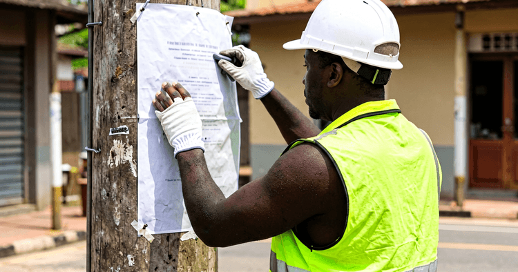 operador eléctrico com capacete, colete reflector e luvas isolantes a trabalhar em poste, de costas para a câmara, sem texto legível