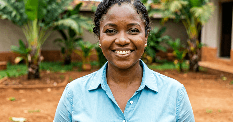 young Mozambican woman in an outdoor courtyard, plain blouse, smiling, chest-up portrait, no legible text
